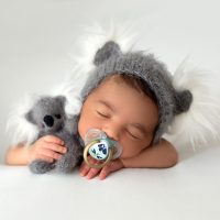 newborn babyboy cute little resting baby with grey hat and grey toy bear in his hand and pacifier on his mouth on a white floor